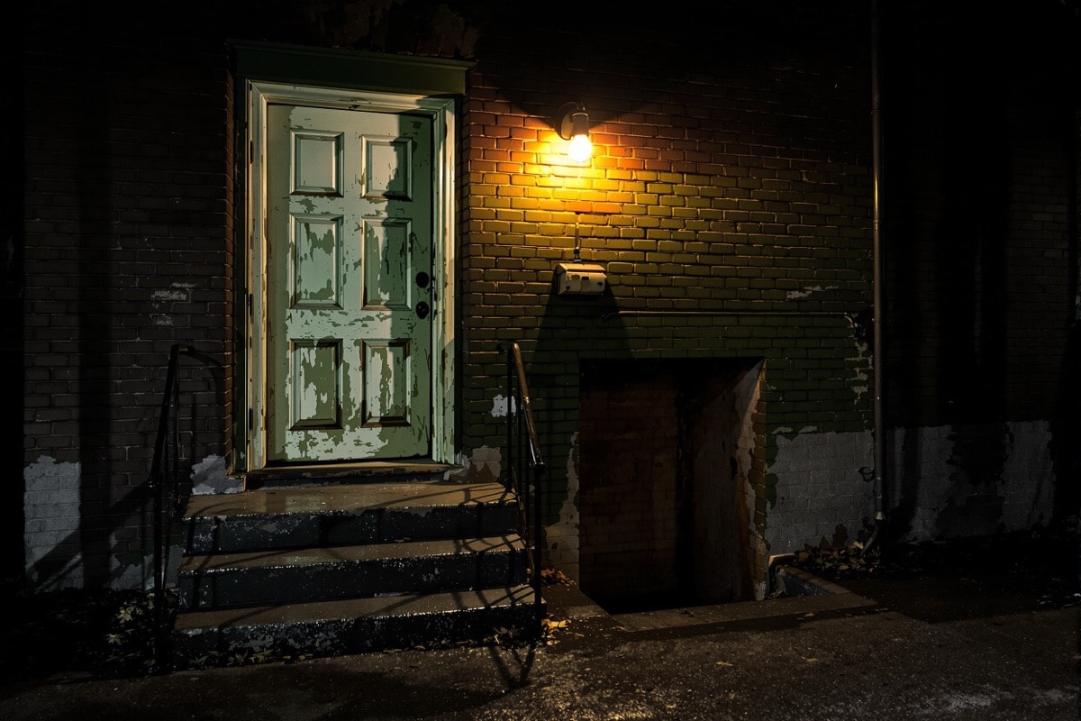 A weathered painted stage door beside a bare bulb, with steps down to a basement entrance
